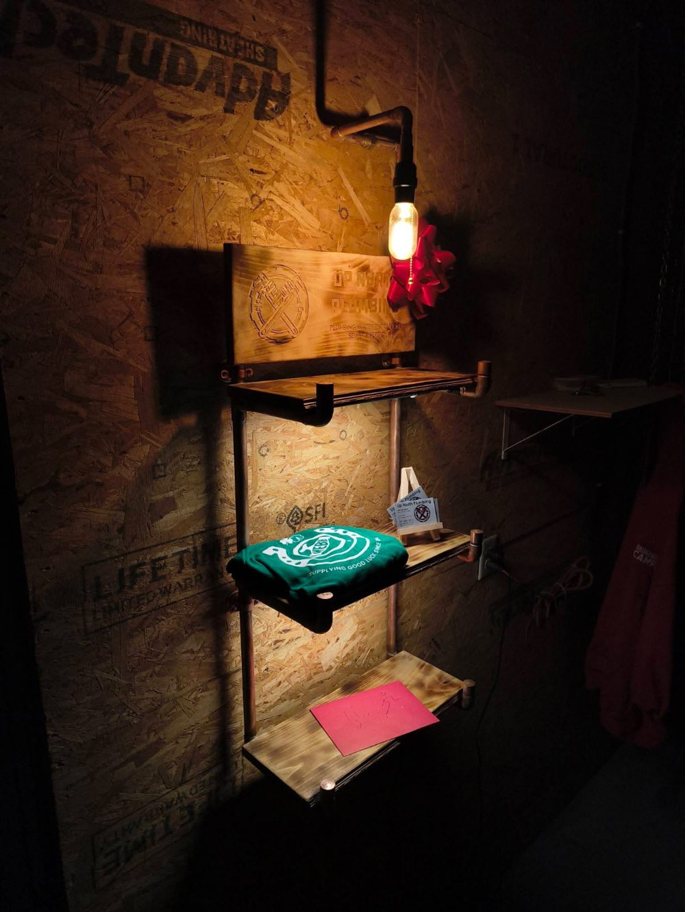 Wall-mounted shelf with copper pipe uprights, torched wood shelves, an engraved sign and an Edison bulb lamp with a pull chain